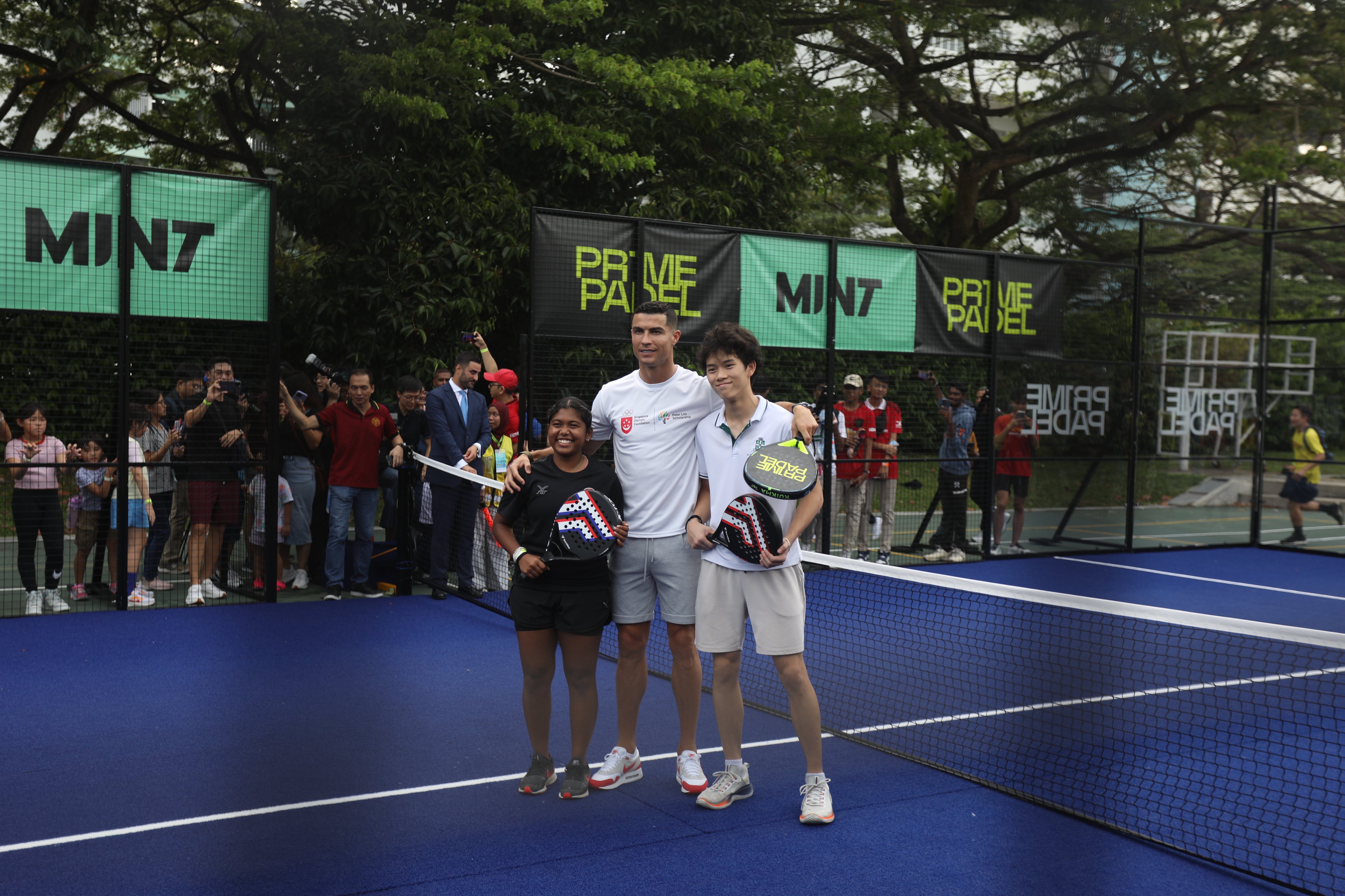 Three people pose with padel rackets on a blue outdoor court, with crowds and Prime Padel banners behind them.